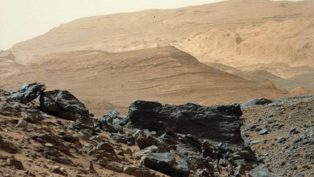 Rocas oscuras en primer plano y estratos del monte Sharp al fondo, fotografiados por el rover Curiosity durante su ascenso en abril de 2015.