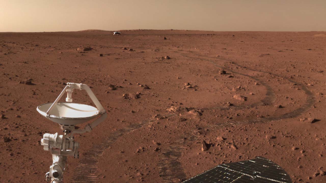 Vista desde el rover Zhurong mostrando sus huellas sobre la superficie de Marte y la etapa de descenso al fondo